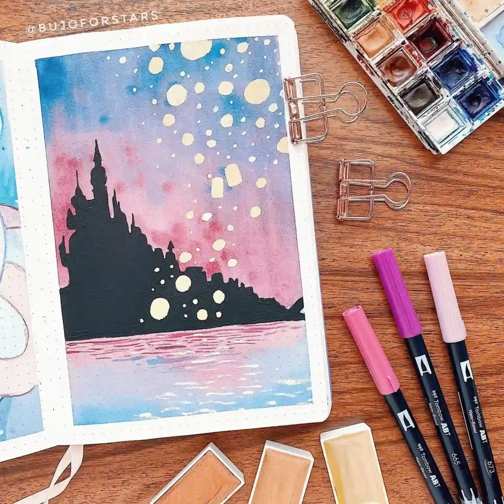Bullet journal art with a starry night castle scene, surrounded by watercolor set and markers on a wooden table.