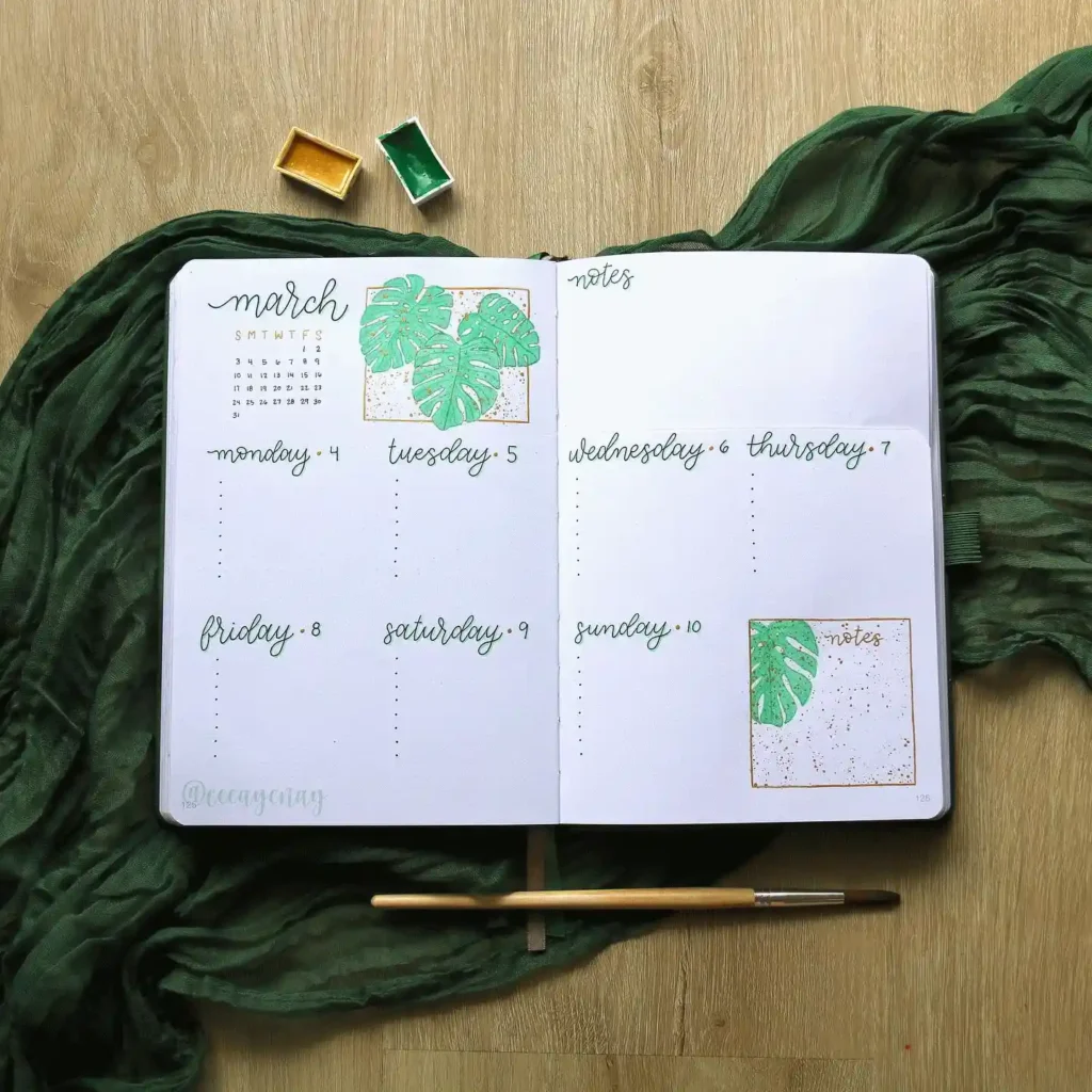 Open journal with weekly planner and green leaf designs on a wooden surface, accompanied by green fabric and a paintbrush.