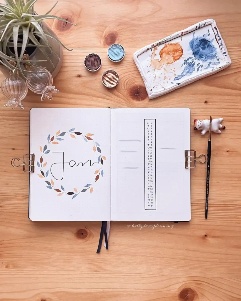 Open bullet journal with artistic January design on wooden desk with paints and plant.