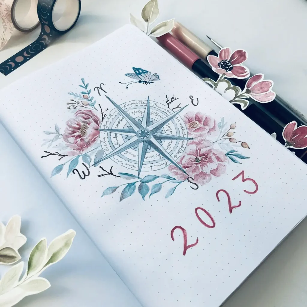 Decorative 2023 bullet journal page with floral compass design, featuring pens and floral cutouts.