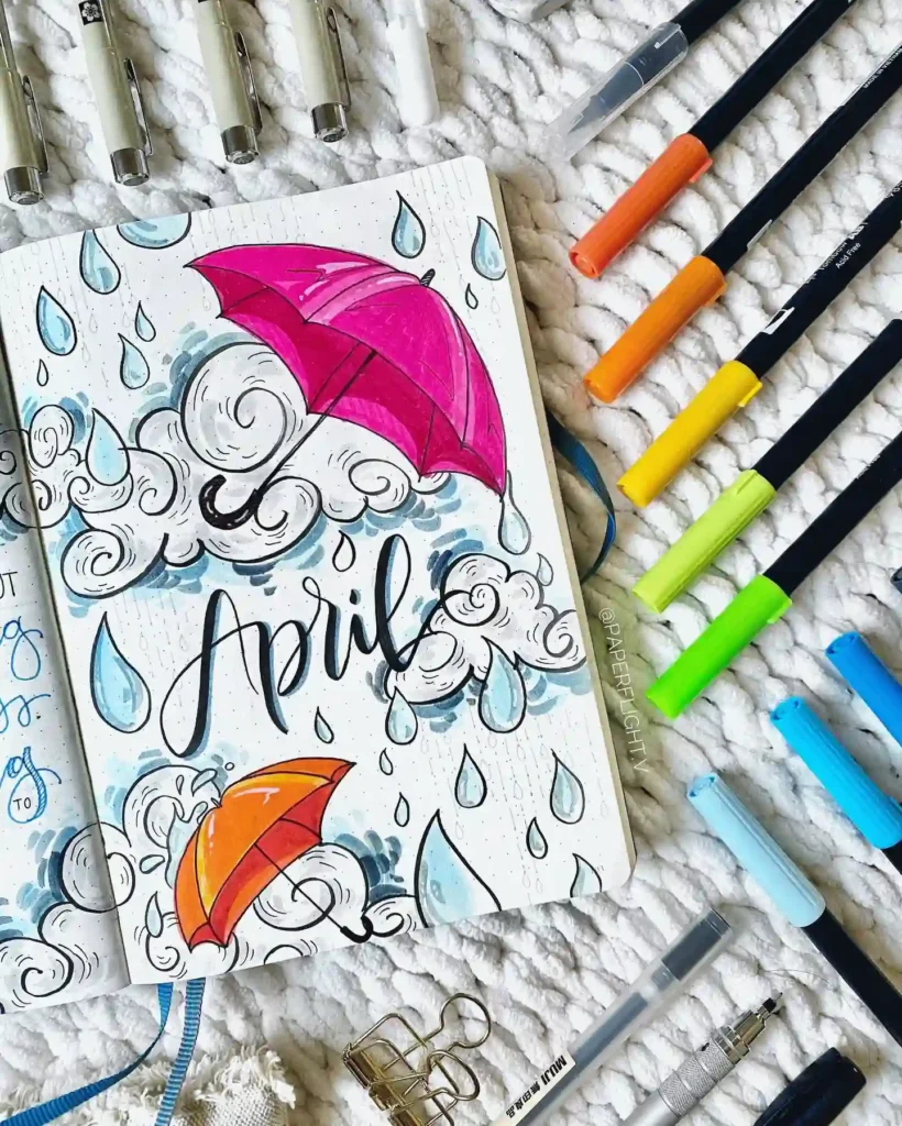 Bullet journal with colorful umbrellas and clouds for April theme, surrounded by markers and stationery.