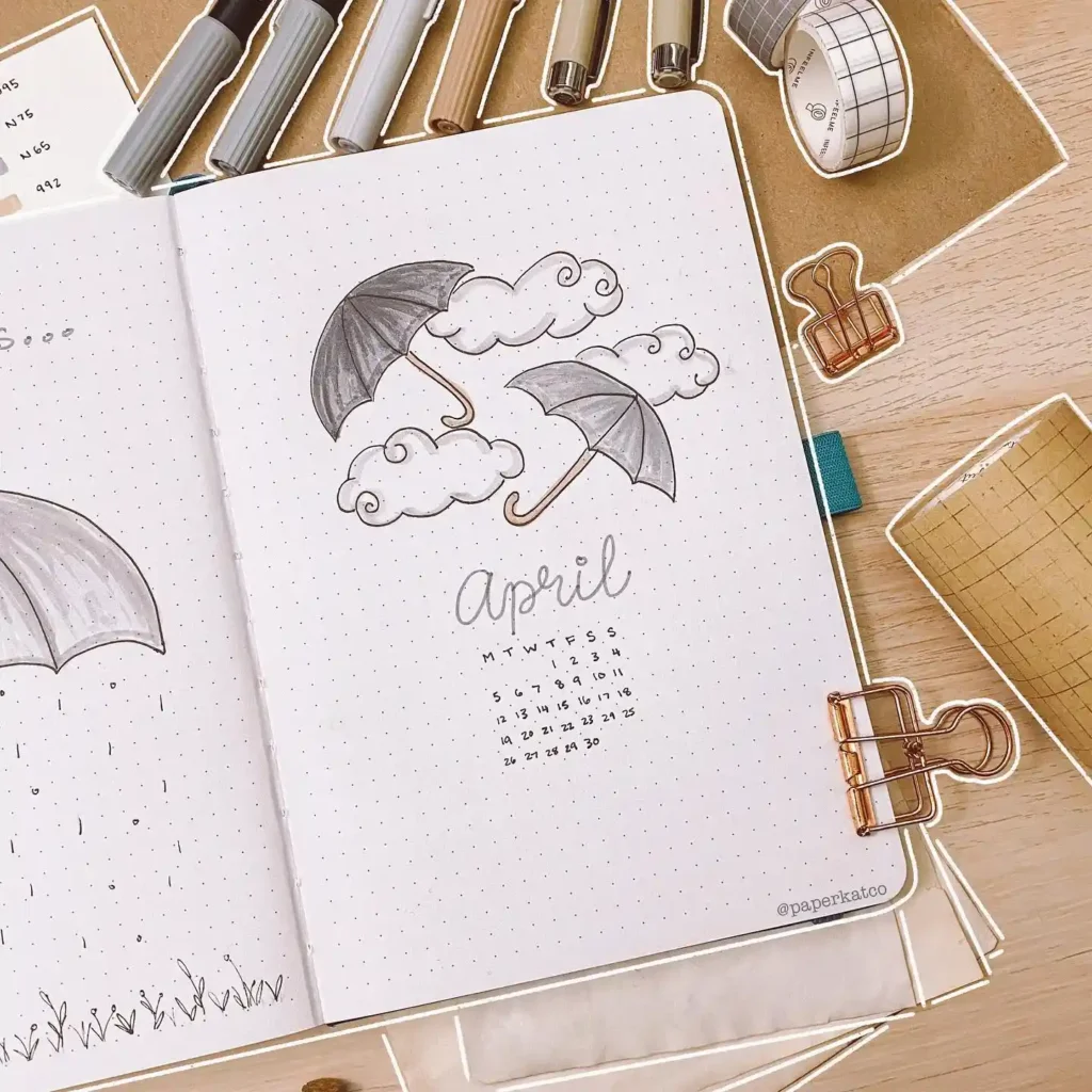 Bullet journal page for April with umbrella doodles and calendar, surrounded by pens and stationery on a desk.