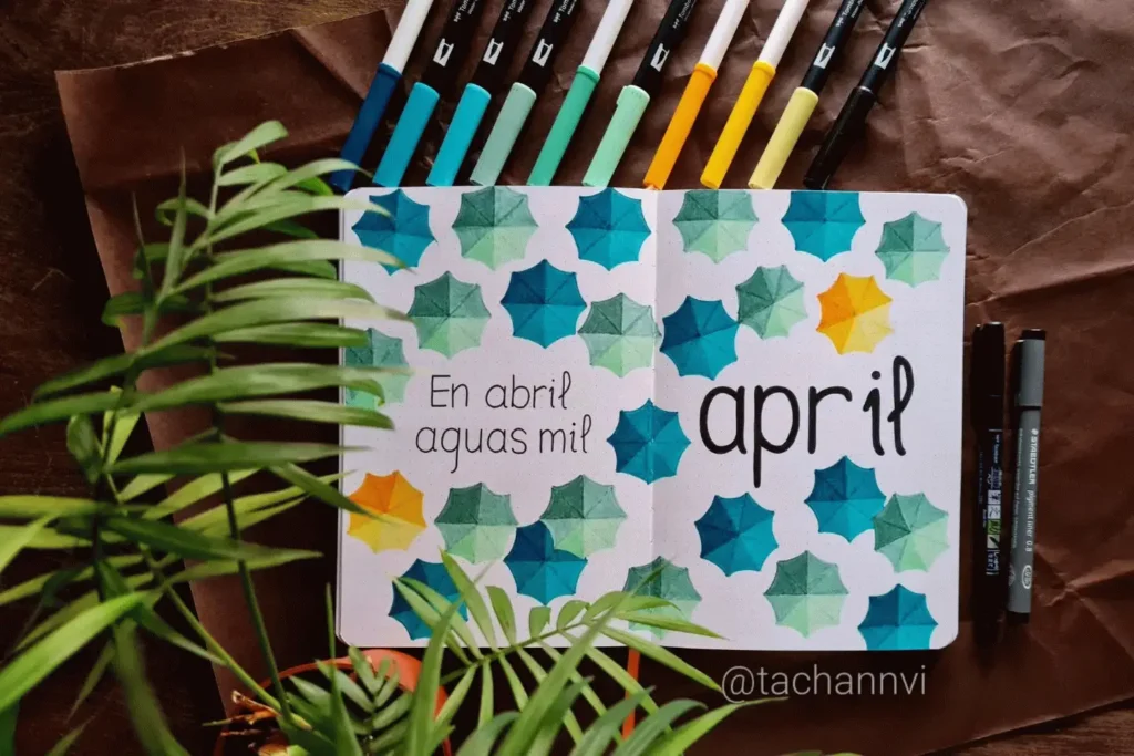 Bullet journal open to April, featuring colorful umbrellas and text, surrounded by pens and a leafy plant.