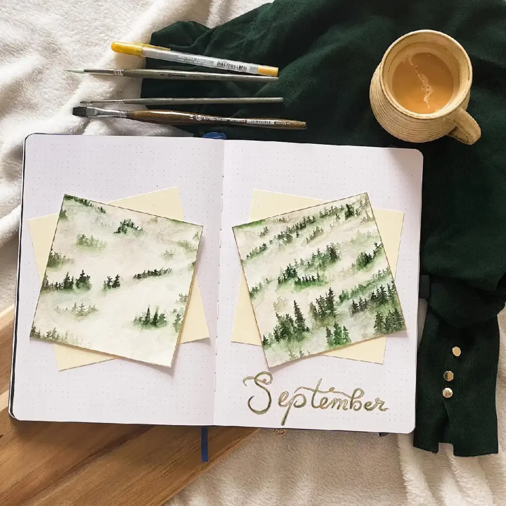 Watercolor paintings of misty forests beside a cozy coffee cup in a bullet journal setup for September.