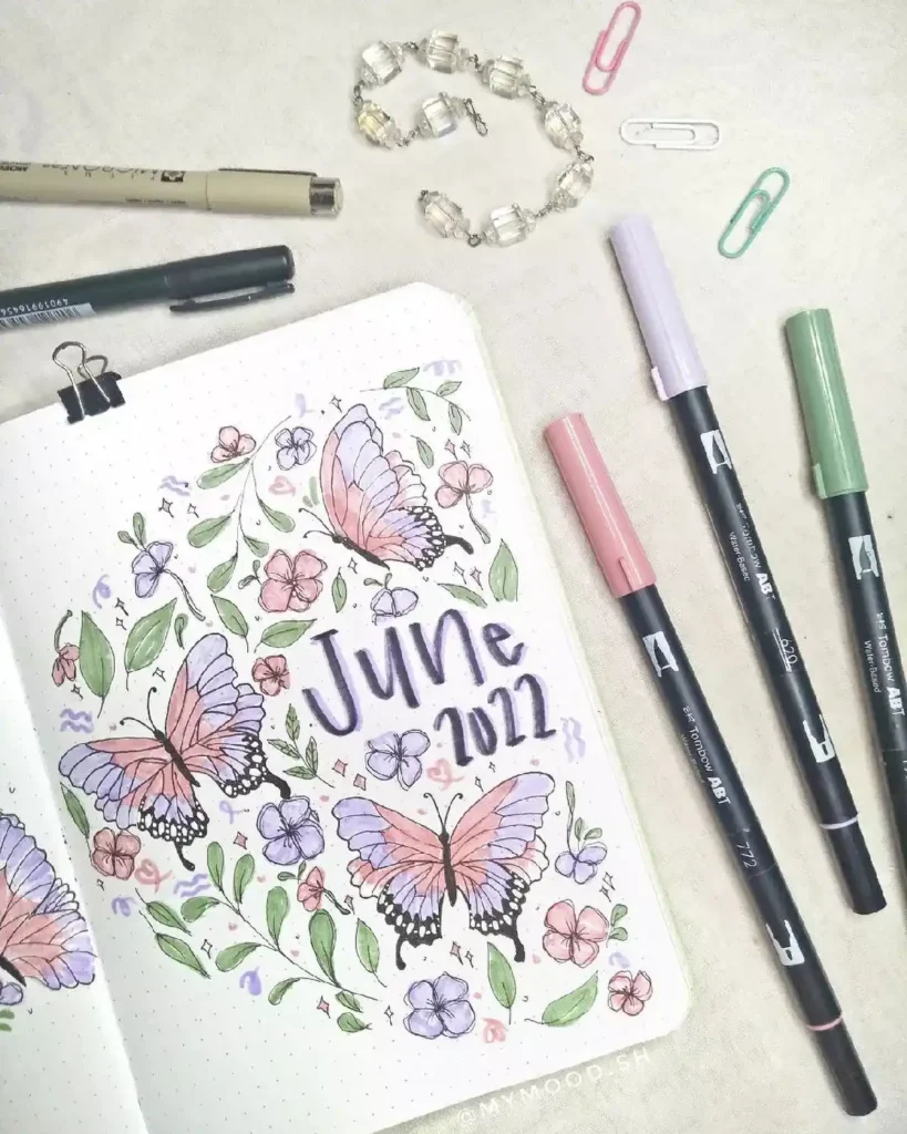 Bullet journal page for June 2022 featuring colorful butterflies and flowers, surrounded by art supplies and paper clips.