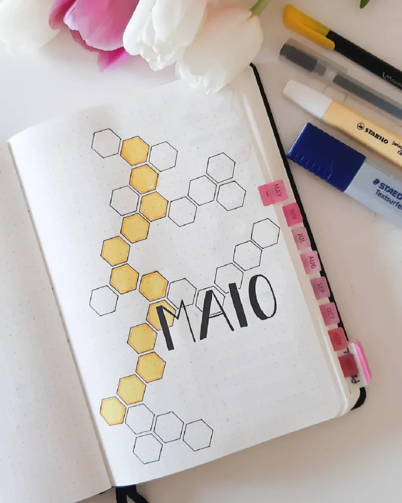 Bullet journal layout featuring honeycomb designs for MAIO with decorative pens and flowers. Perfect for planning.