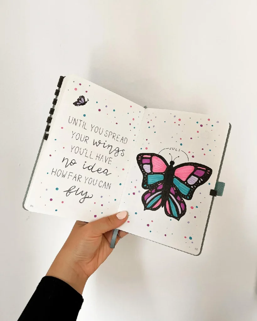 Hand holding an open notebook featuring a colorful butterfly illustration and an inspiring quote about spreading wings.