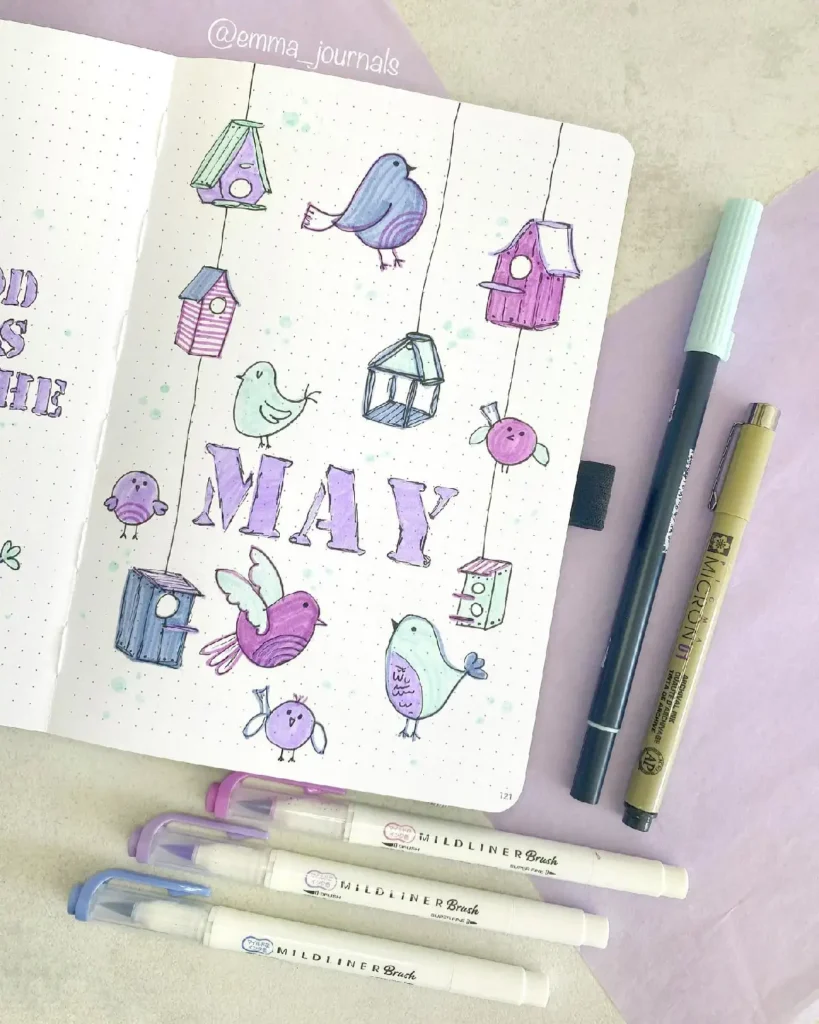 Colorful bullet journal page for May, featuring birds and birdhouses, alongside various pens for creative planning.