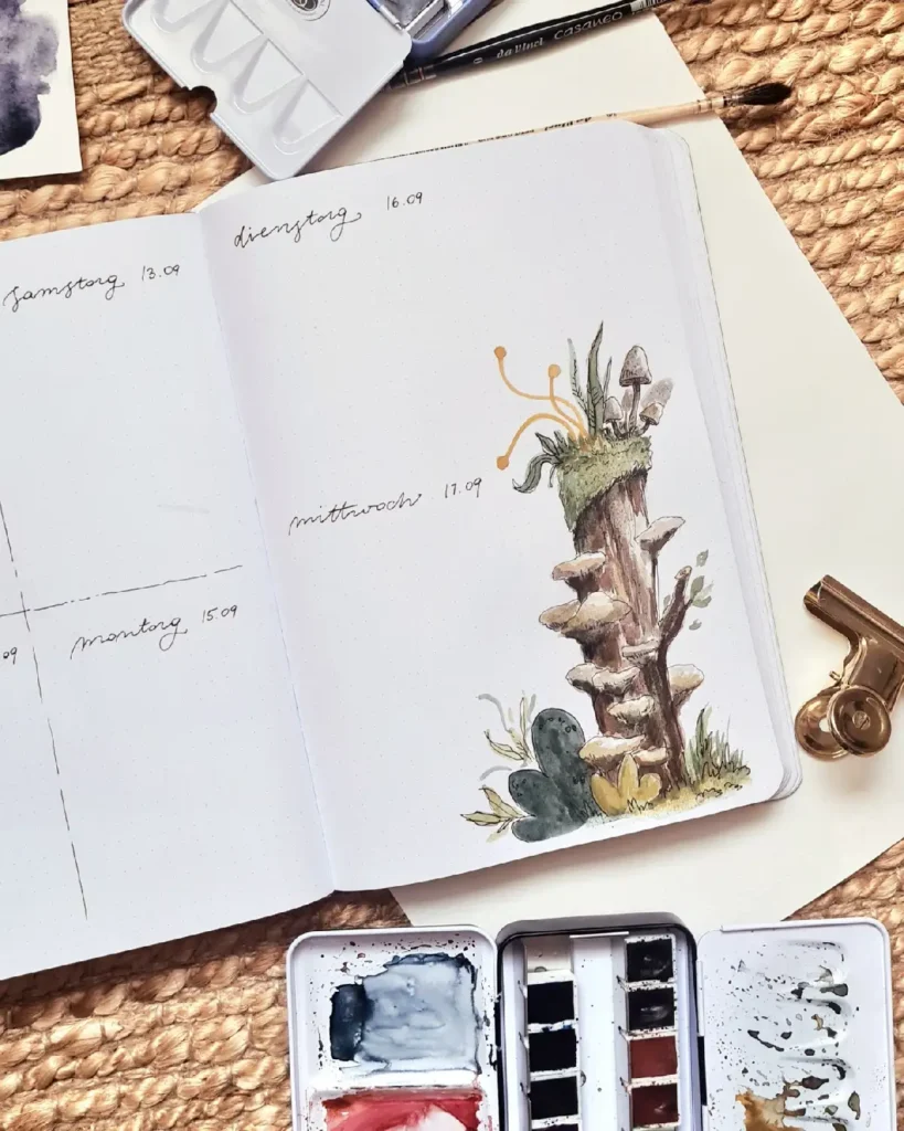 Open watercolor journal with a mushroom illustration, paint palette, and stationery on a textured background.