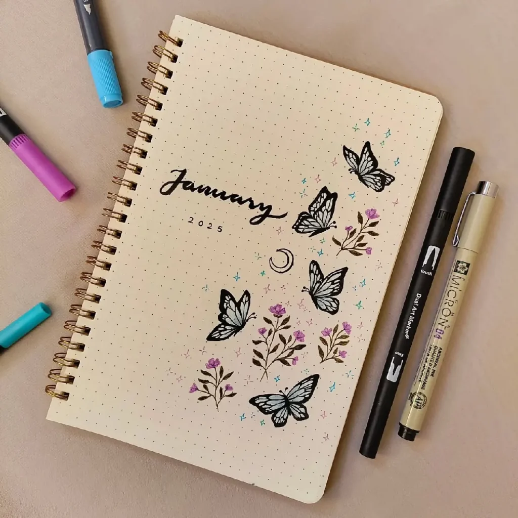 Stylish bullet journal for January 2025, decorated with butterflies and flowers, surrounded by colorful pens.