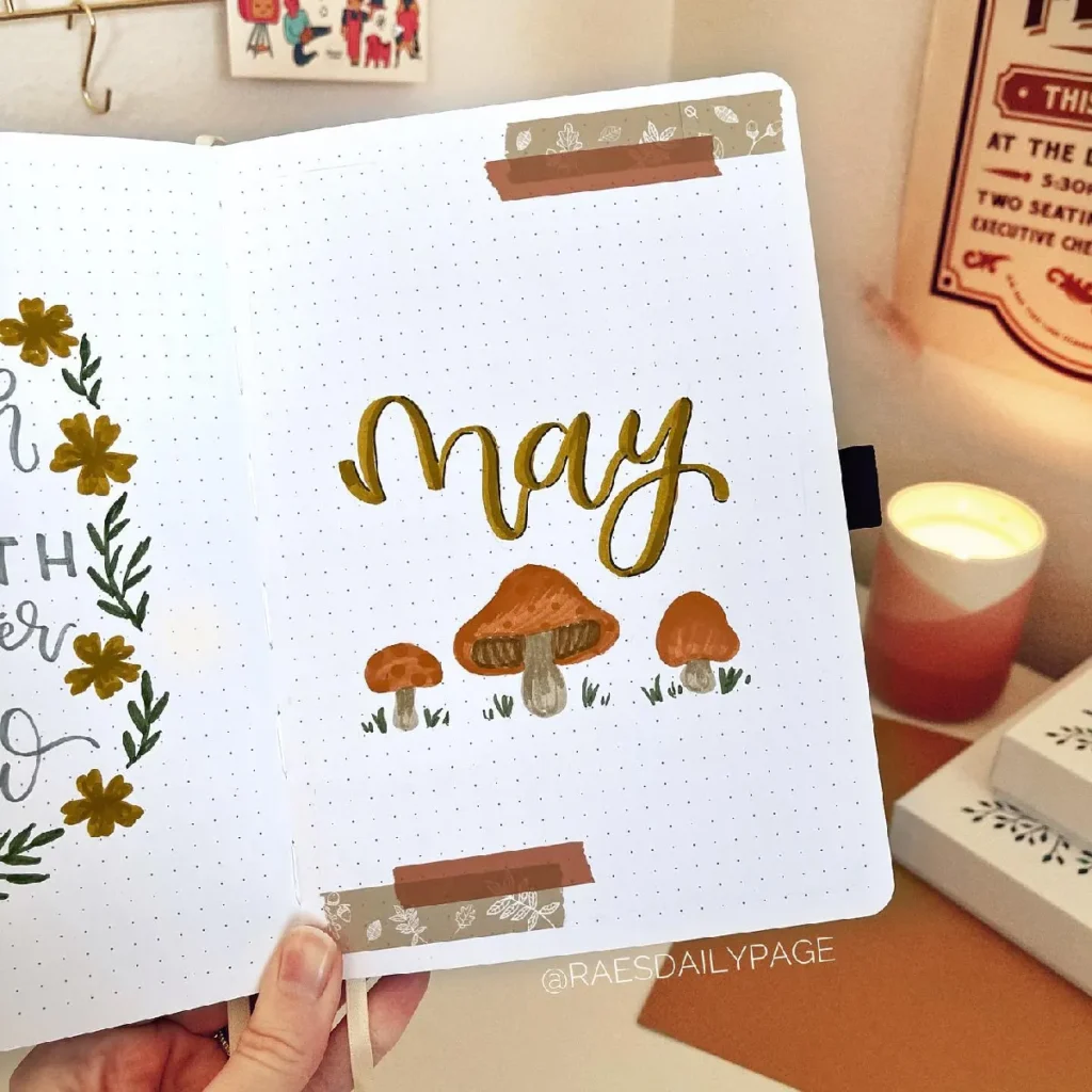 Handwritten May on dotted journal page with mushrooms, flowers, and decorative tape, featuring a cozy candle in the background.