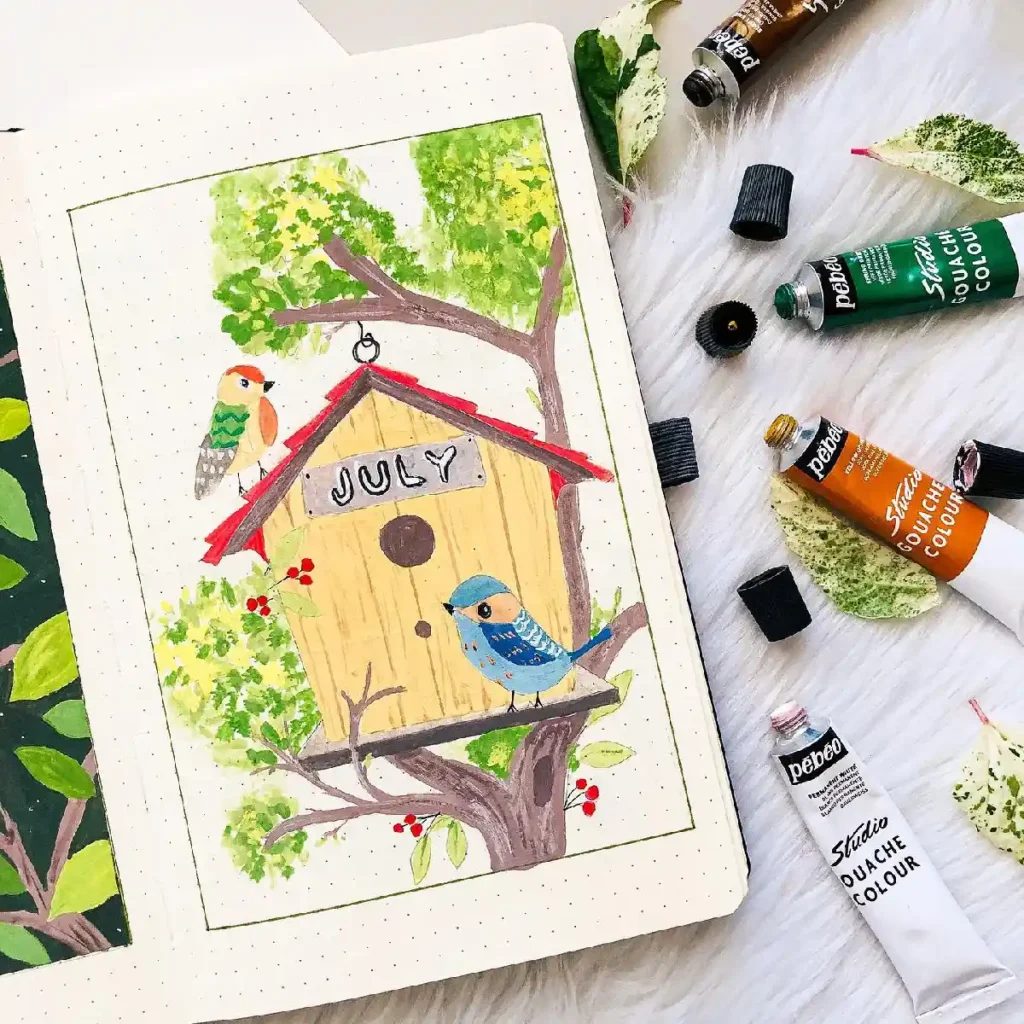 A bullet journal page for July featuring a colorful birdhouse and two birds, with art supplies nearby.